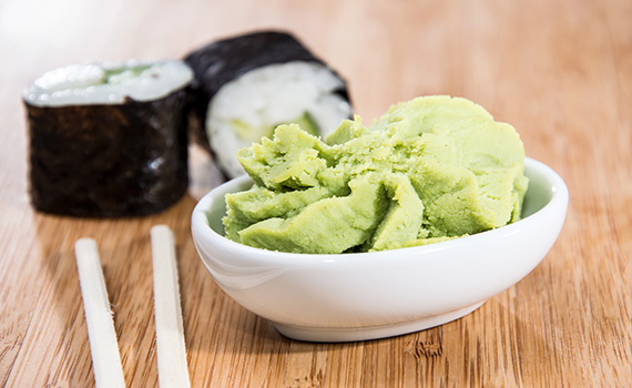 Wasabi preparado numa tigela branca, dois pauzinhos ao lado e dois rolos de sushi atrás. 