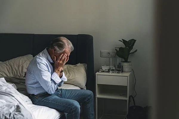 Um idoso está sentado na beira da cama, segurando a cabeça devido à dor.