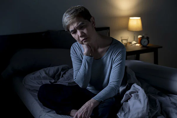 Uma mulher idosa sentada na beira da cama, com expressão de tristeza e exaustão, refletindo os efeitos da insônia e da depressão.