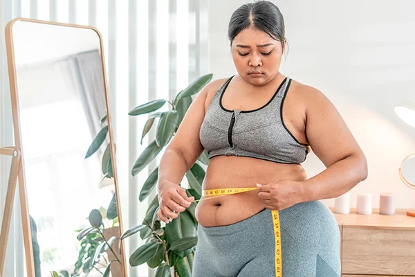 Uma mulher com excesso de peso mede-se com uma fita métrica em frente ao espelho. 
