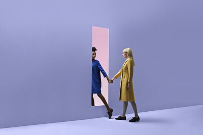 two-women-holding-hands-walking-threw-rectangular-opening-in-coloured-wall