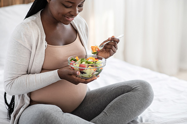 Mujer embarazada sentada en su cama mientras come una ensalada.