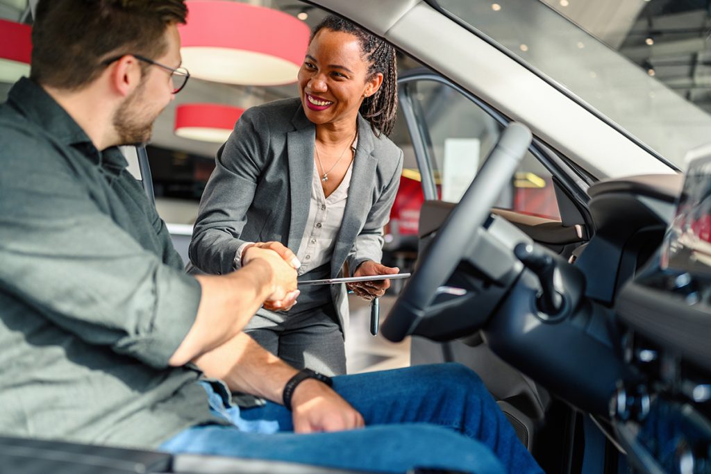 Hombre se decide a comprar un auto, así que, sentado en el asiento de piloto y con la puerta abierta, estrecha la mano de la vendedora en señal de haber llegado a un acuerdo. 