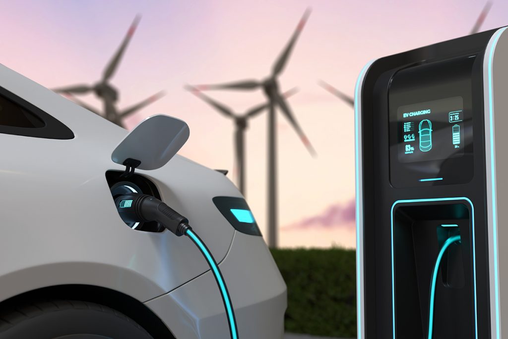 Carro eléctrico cargándose en una estación de carga. Al fondo una granja de energía eólica, representando que ambas son fuente de energía limpia.