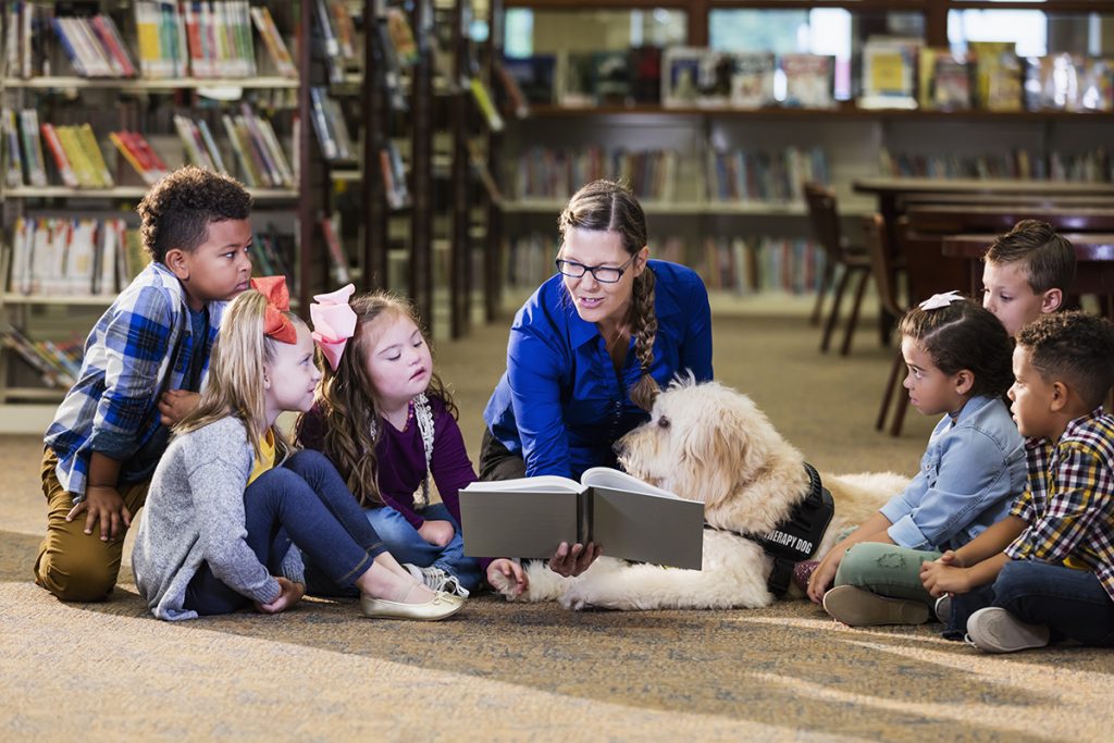 Sentados sobre el suelo de la biblioteca, una muestra y sus estudiantes leen un libro, acompañado por un perro de terapia.
