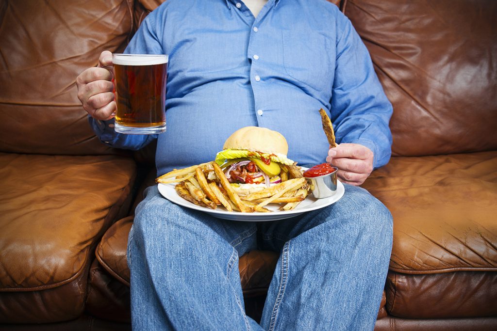 Persona con obesidad sentado en un sillón sostiene una cerveza y come una hamburguesa con papas fritas.