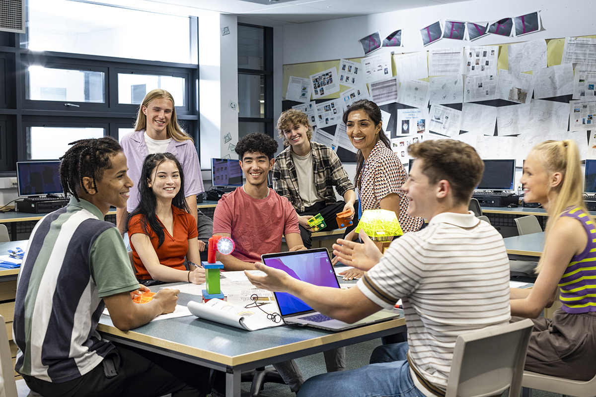 Gran plano de un grupo de estudiantes y su profesor reunidos alrededor de una mesa en un aula de tecnología.